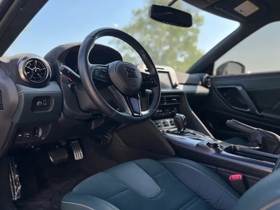 Interior view of a Nissan GT-R, showing the steering wheel, dashboard, center console with carbon fiber trim, and pedals.