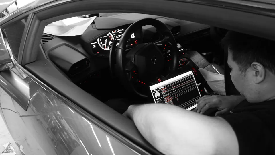 A person tuning a Lamborghini's ECU with a laptop in a shop, showing the car's dashboard and steering wheel.