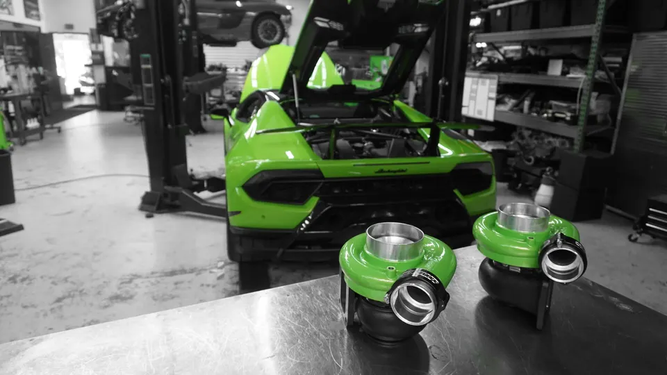 Green Lamborghini Huracan on a lift in a performance shop, with two green turbochargers on a workbench in the foreground.