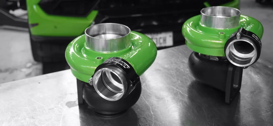 Two bright green turbochargers on a metal table in a performance shop, with a green Lamborghini in the background.