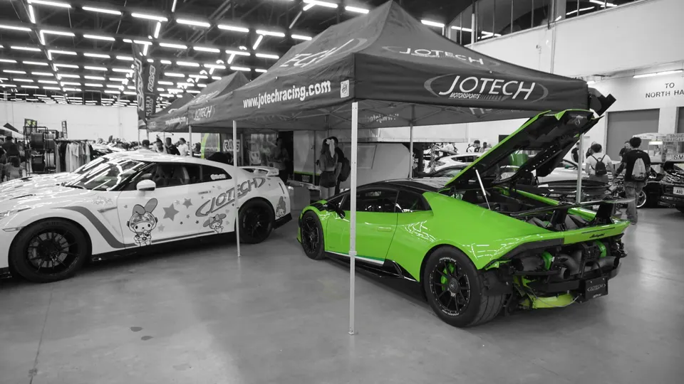 A green Lamborghini Huracan with exposed rear turbos and a white Nissan GT-R with custom livery under Jotech Motorsports tents at a car show.