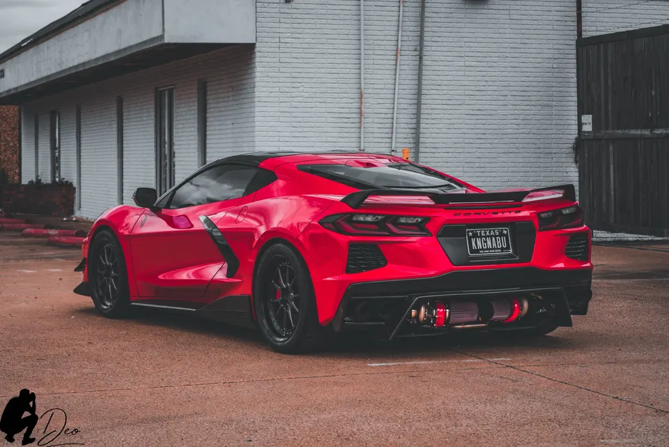 A red C8 Corvette with a visible twin-turbo setup and custom rear diffuser parked in front of a white brick building.