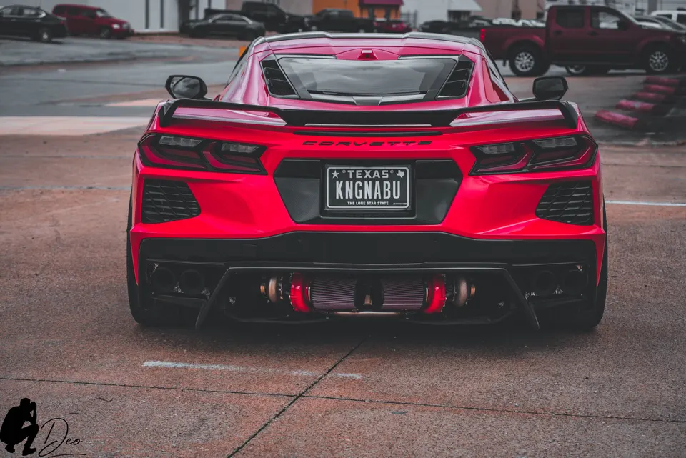 Red C8 Corvette with exposed twin turbochargers and custom exhaust, parked outdoors. Texas license plate "KNGNABU".