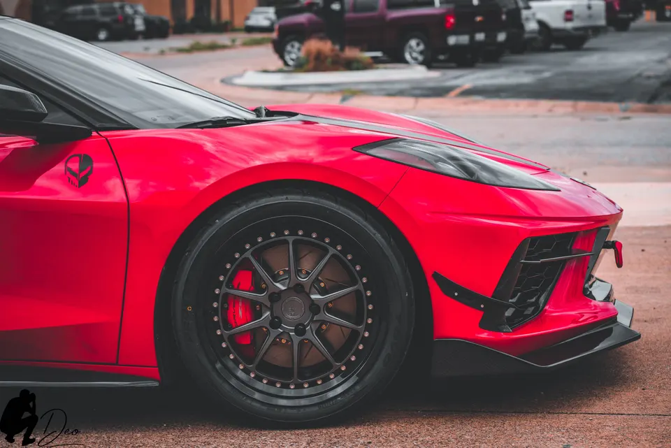 A close-up detail shot of a red C8 Corvette featuring custom black multi-spoke wheels, red brake calipers, and a black front splitter.
