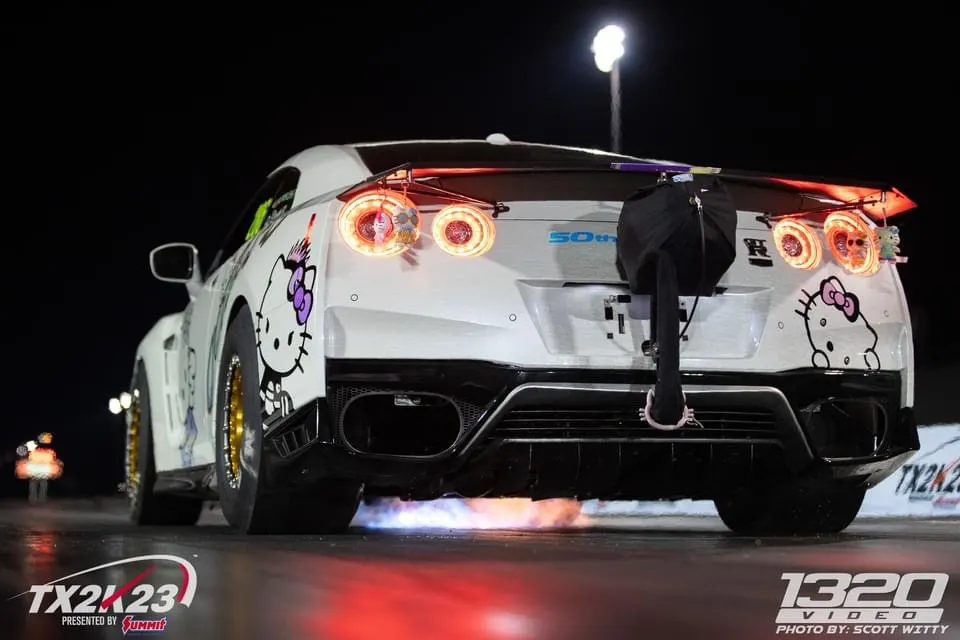 White Nissan GT-R with Hello Kitty decals launching on a drag strip at night, with flames shooting from the exhaust.
