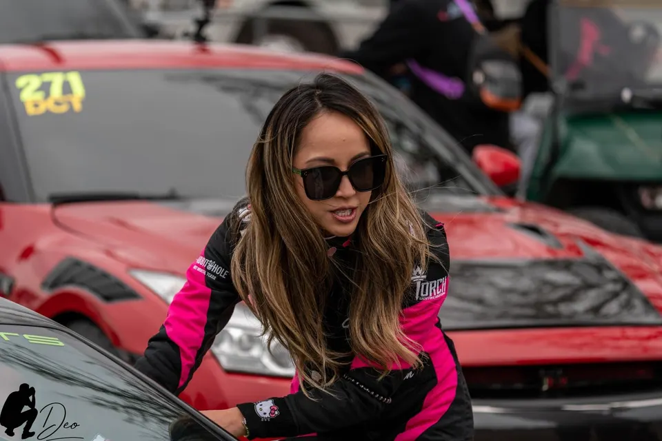 A woman in a pink and black racing suit and sunglasses leans on a car at a drag racing event, with a red Nissan GT-R in the background.