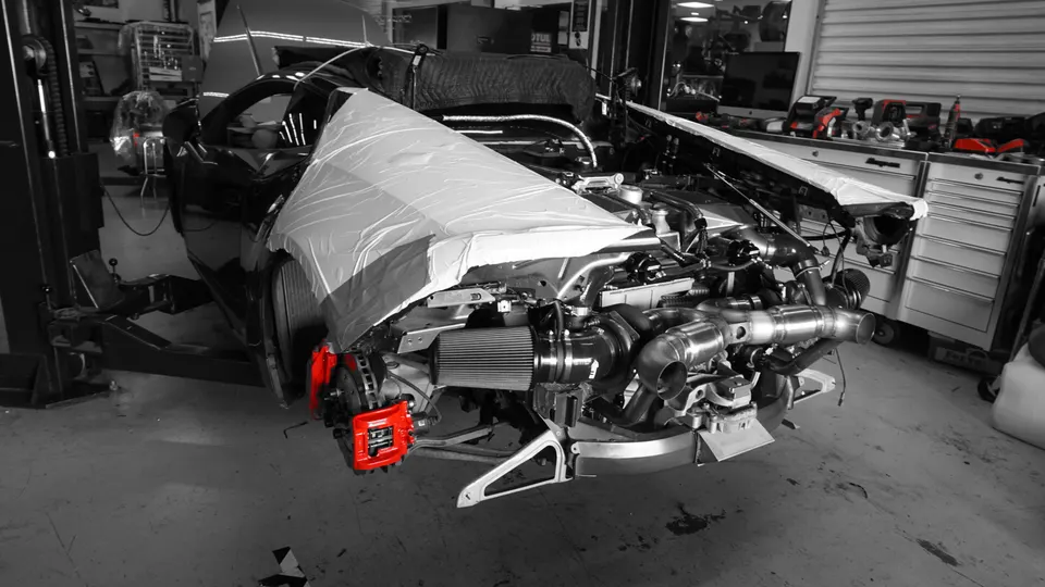 Car in a shop with its front disassembled, revealing a large turbocharger, air filter, and red brake calipers during a performance build.
