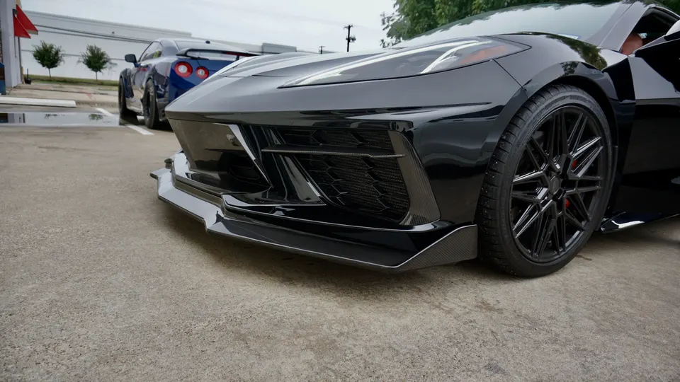 Black Chevrolet Corvette C8 with a carbon fiber front splitter and aftermarket wheels, parked with a blue Nissan GT-R.