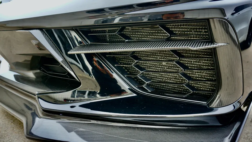 Close-up of a car's front bumper featuring a carbon fiber splitter and grille with honeycomb mesh.