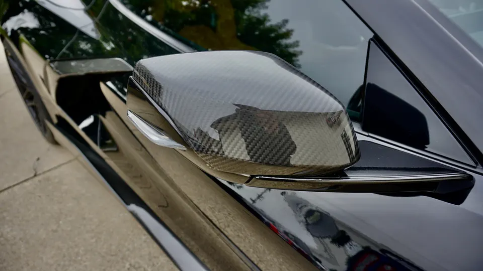 Close-up of a black car's carbon fiber side mirror, showcasing the weave pattern and glossy finish.
