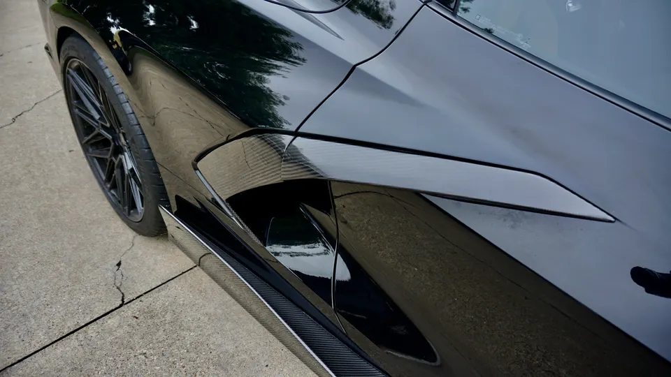 A close-up detail shot of a glossy black sports car's side, showing a carbon fiber side skirt, rear wheel, and body lines.