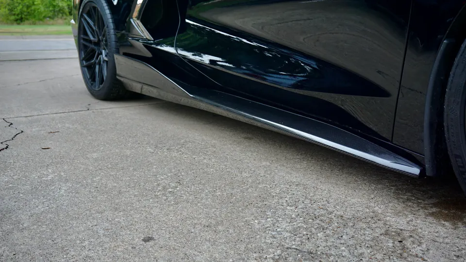 Close-up of a black car's carbon fiber side skirt on concrete, highlighting the aerodynamic detail.
