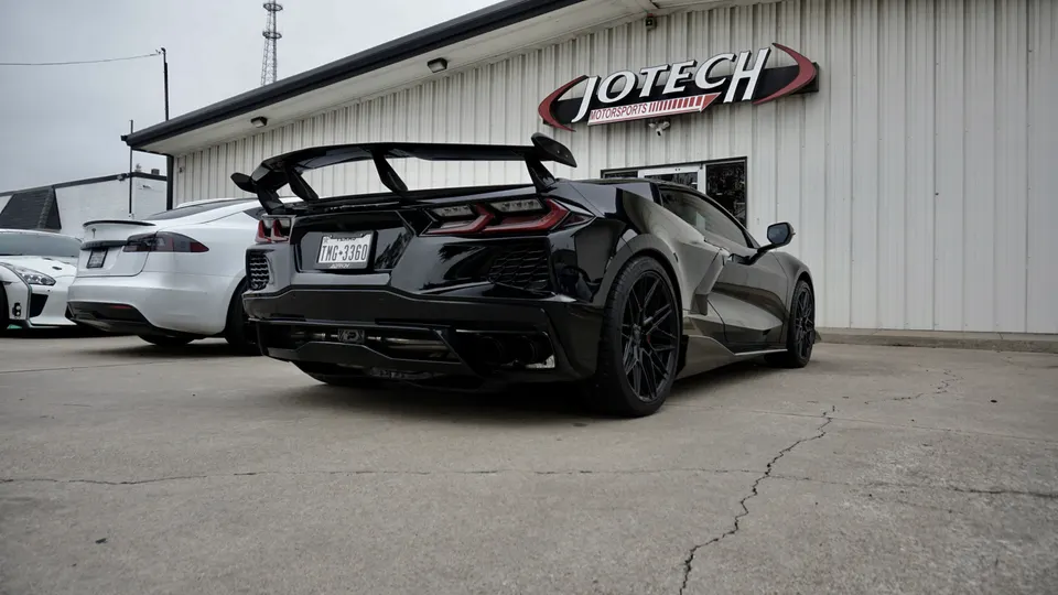 A black Corvette C8 with a large rear wing parked outside Jotech Motorsports, with other cars in the background.