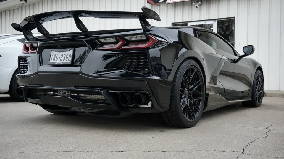 Black C8 Corvette with large aftermarket rear wing, custom wheels, and performance exhaust system parked outside.