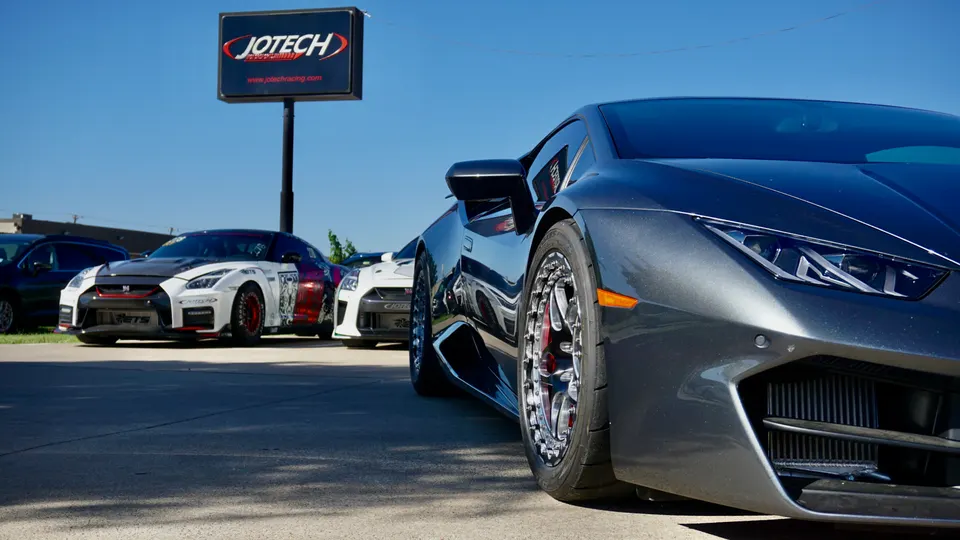 Modified Lamborghini Huracan and Nissan GT-R parked outside Jotech Motorsports performance tuning shop under a clear sky.