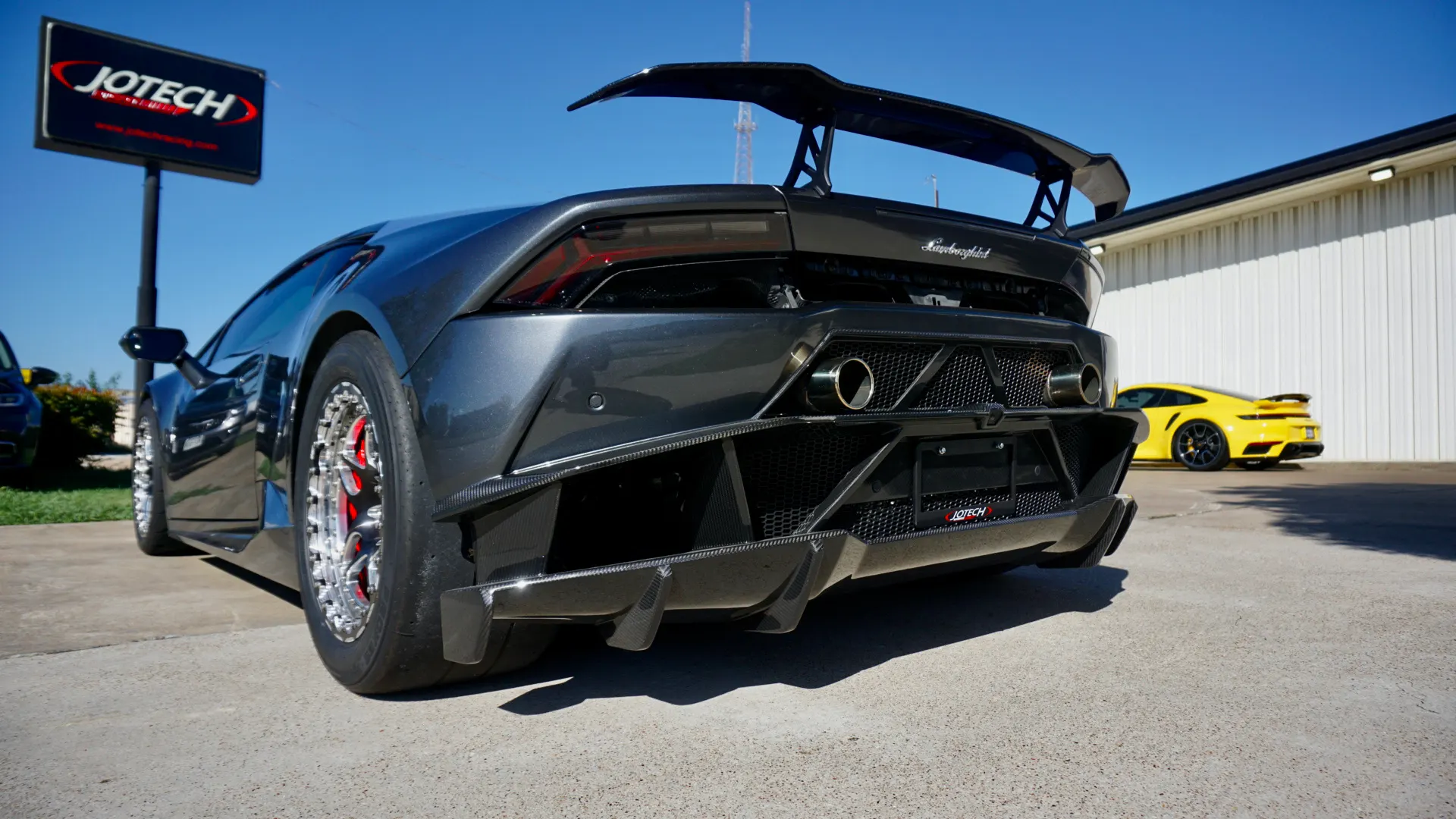 Rear view of a modified dark grey Lamborghini Huracan with a large carbon fiber wing and diffuser, parked outside Jotech Motorsports.