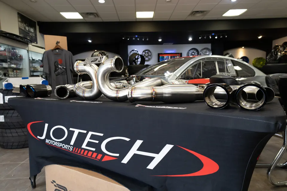 Jotech Motorsports shop display featuring a high-performance exhaust system with polished downpipes and black exhaust tips.