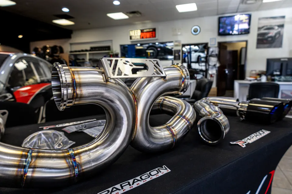 Stainless steel performance exhaust system with downpipes and muffler tips displayed on a table at a tuning shop.