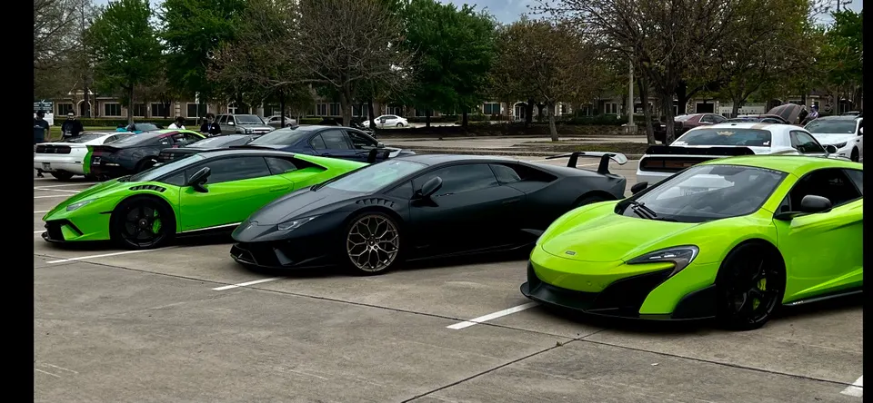 A line of exotic supercars including a green Lamborghini Huracan, a black Lamborghini, and a green McLaren parked at a car meet.