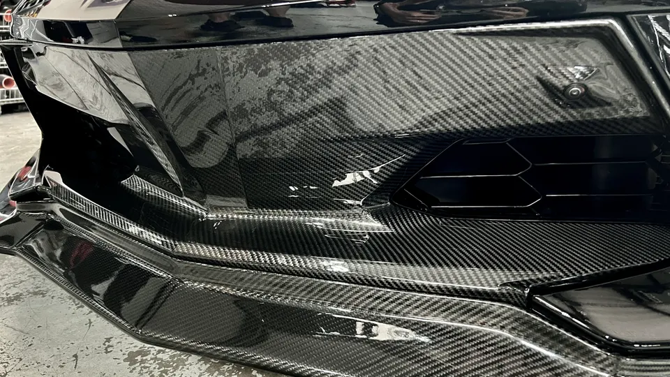 A close-up of a car's carbon fiber front bumper and splitter, showcasing the weave pattern and glossy finish.