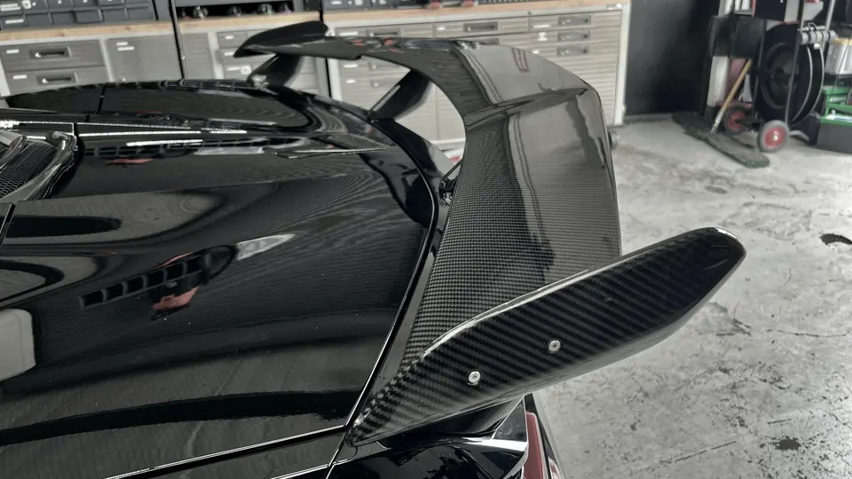 Close-up of a black car's carbon fiber rear wing in a shop environment.