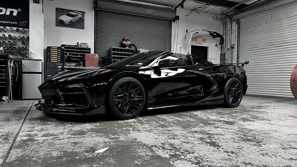 A black C8 Corvette convertible with custom wheels and aftermarket aero parked inside a performance tuning shop.