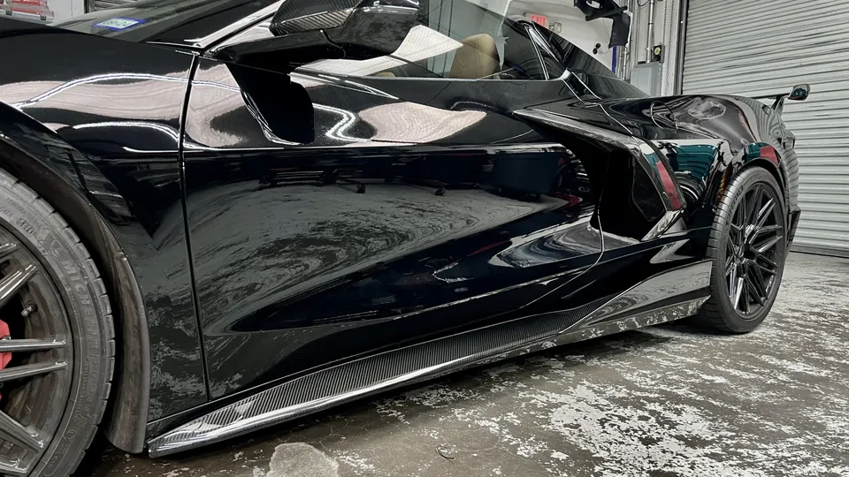 Black C8 Corvette with carbon fiber side skirts and black multi-spoke wheels in a garage setting.