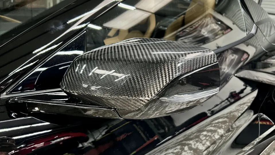 Close-up of a carbon fiber side mirror on a black sports car, showcasing the weave pattern and glossy finish.