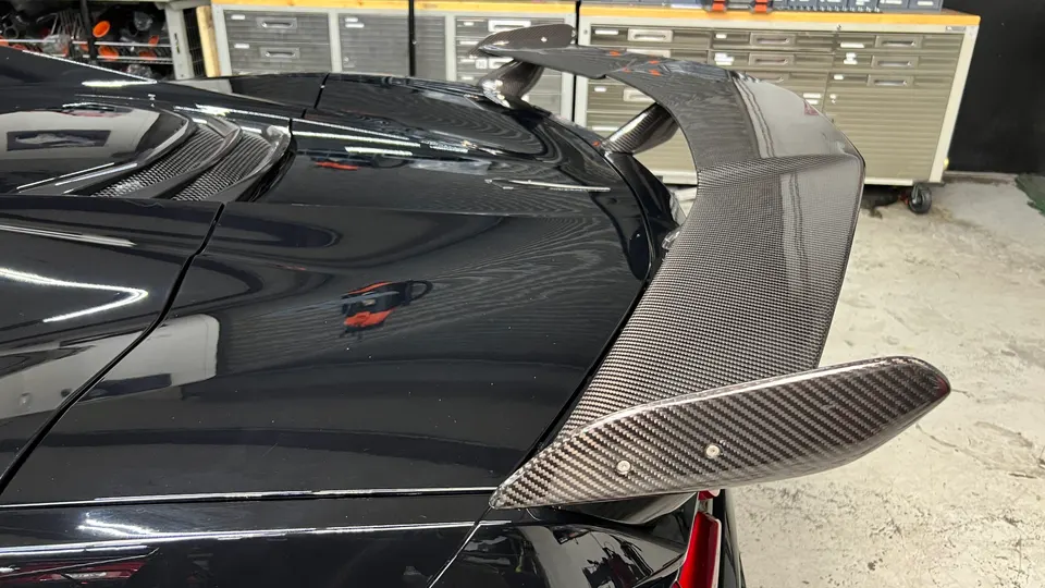 Close-up of a black car's rear with a large carbon fiber wing, showcasing the weave pattern in a shop setting.