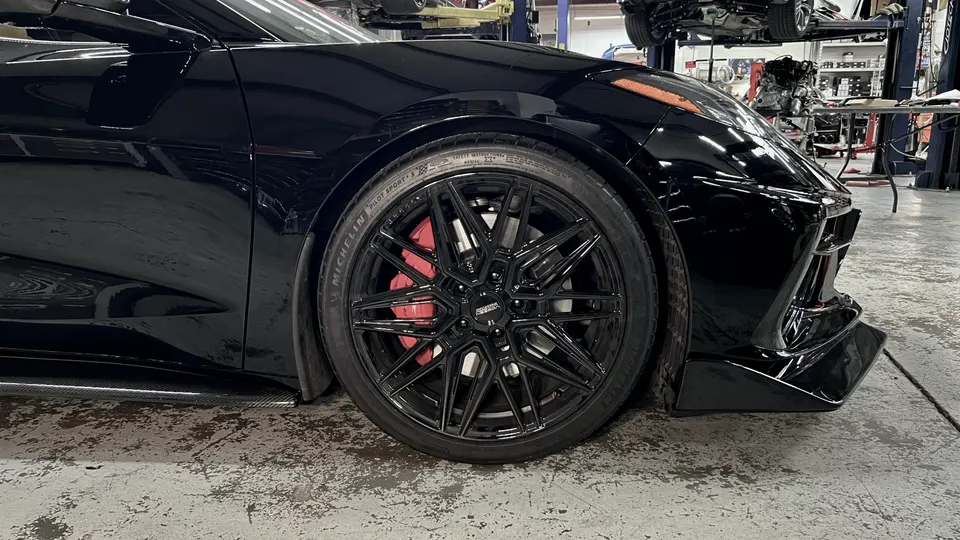 Black Corvette C8 with aftermarket black wheels, red brake calipers, and carbon fiber aero in a performance shop.
