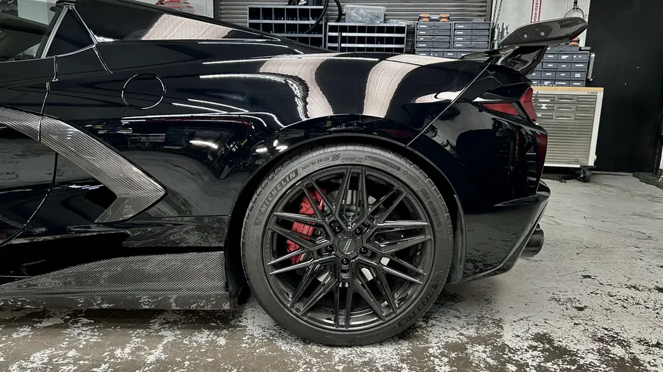 A black Corvette with carbon fiber aerodynamic parts, aftermarket black wheels, and red brake calipers in a shop.