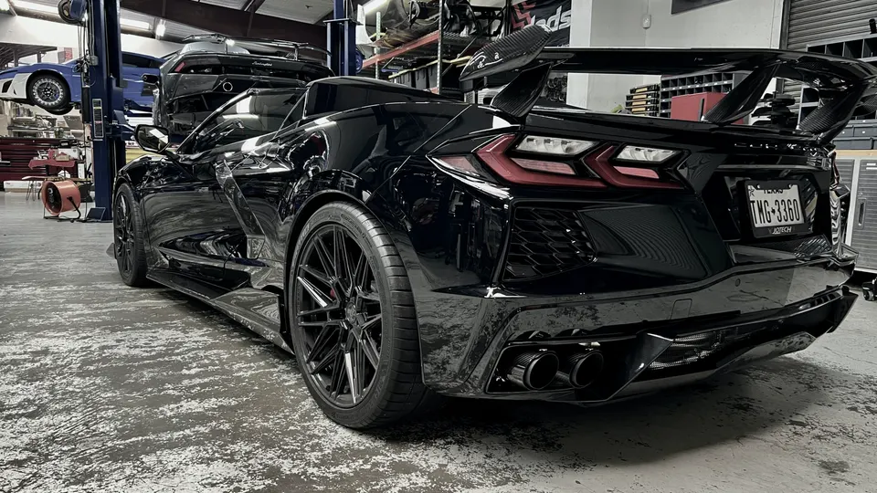A black C8 Corvette with a large carbon fiber wing and aftermarket wheels parked in a performance tuning shop.