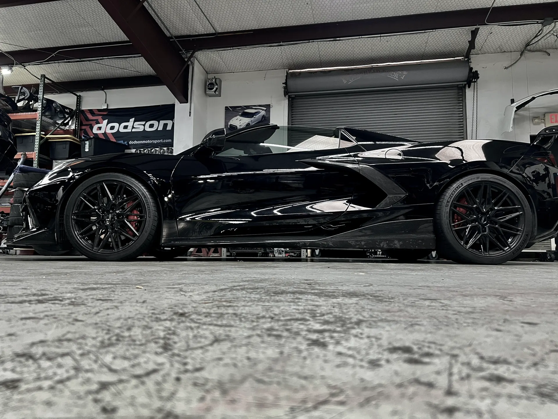A black C8 Corvette with aftermarket wheels and aero modifications parked inside a performance tuning shop.