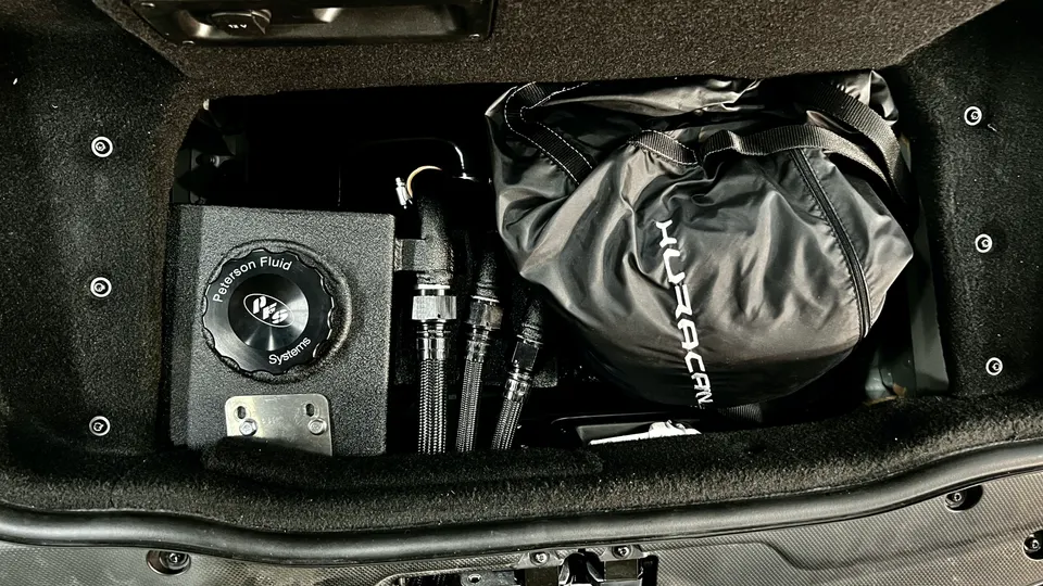 Peterson Fluid Systems tank and braided lines installed in the trunk of a performance car, next to a black bag.