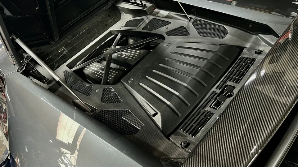 Close-up view of a Lamborghini engine bay featuring a black engine cover with Lamborghini branding and carbon fiber trim.