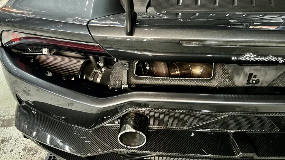 Rear view of a grey Lamborghini with exposed custom turbocharger, air filter, and exhaust system, featuring carbon fiber trim.