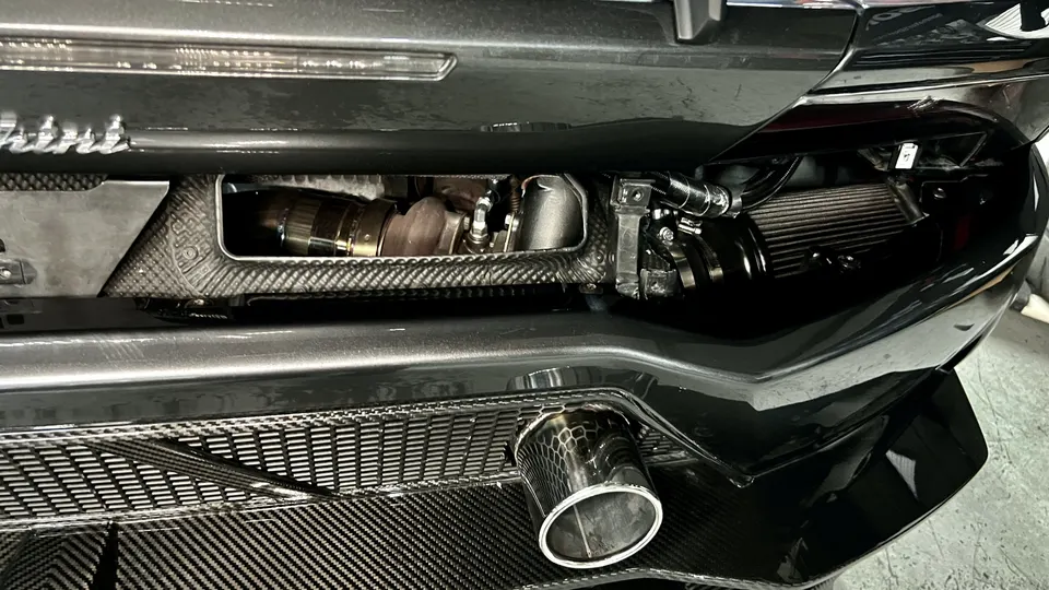 Close-up of a performance car's rear bumper, showcasing carbon fiber, an exposed turbocharger, intercooler, and exhaust tip.