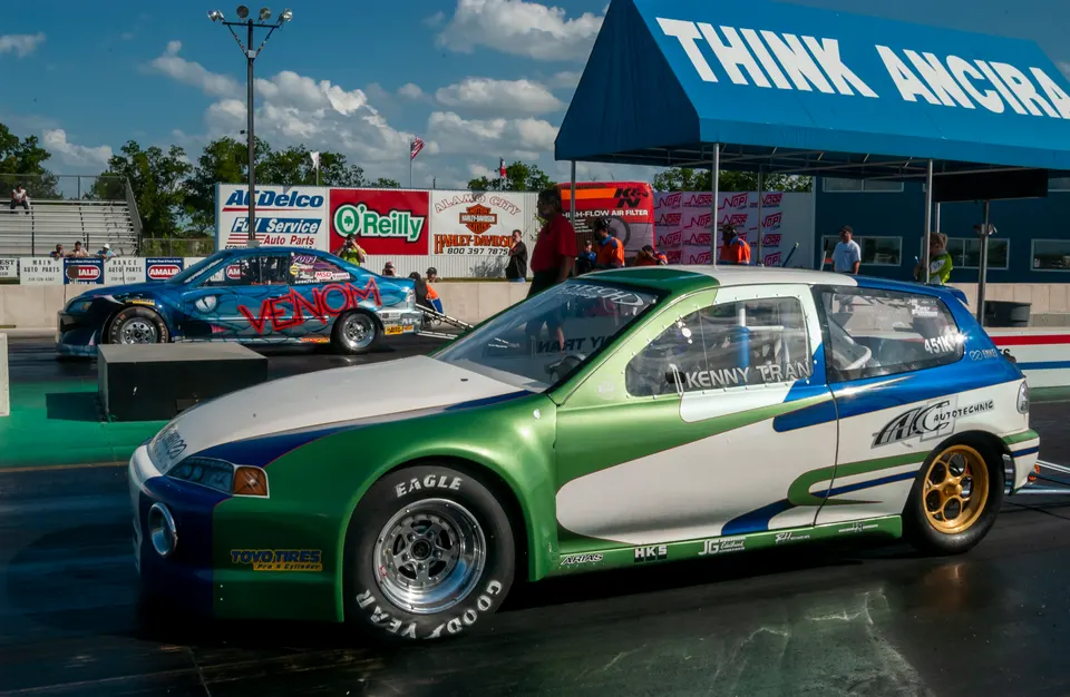 Two custom-painted drag racing cars, a green, white, and blue Honda Civic and a blue "Venom" car, at the starting line of a drag strip.