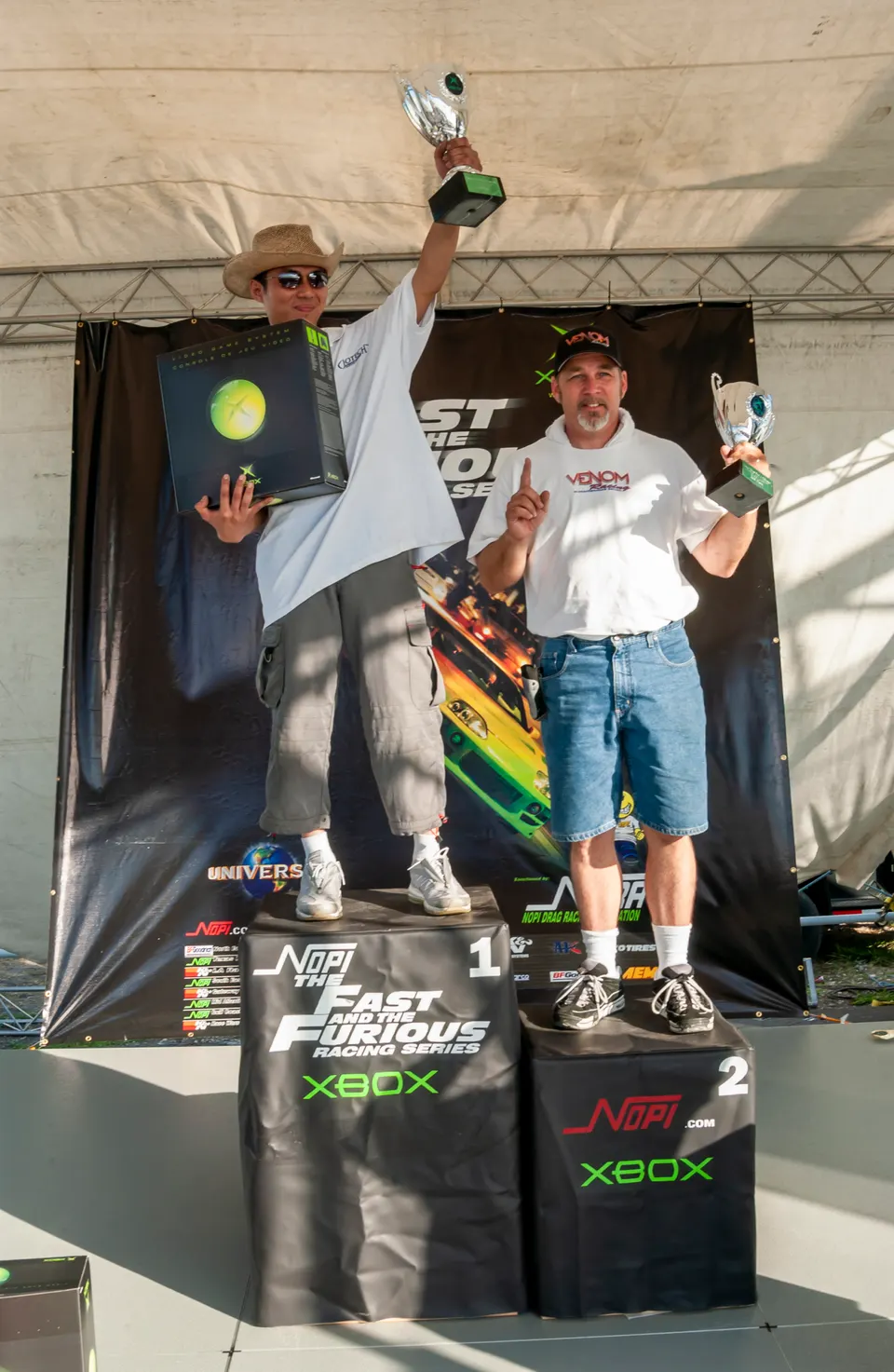 Two winners on a podium holding trophies and an Xbox console at a Fast and Furious Racing Series event.