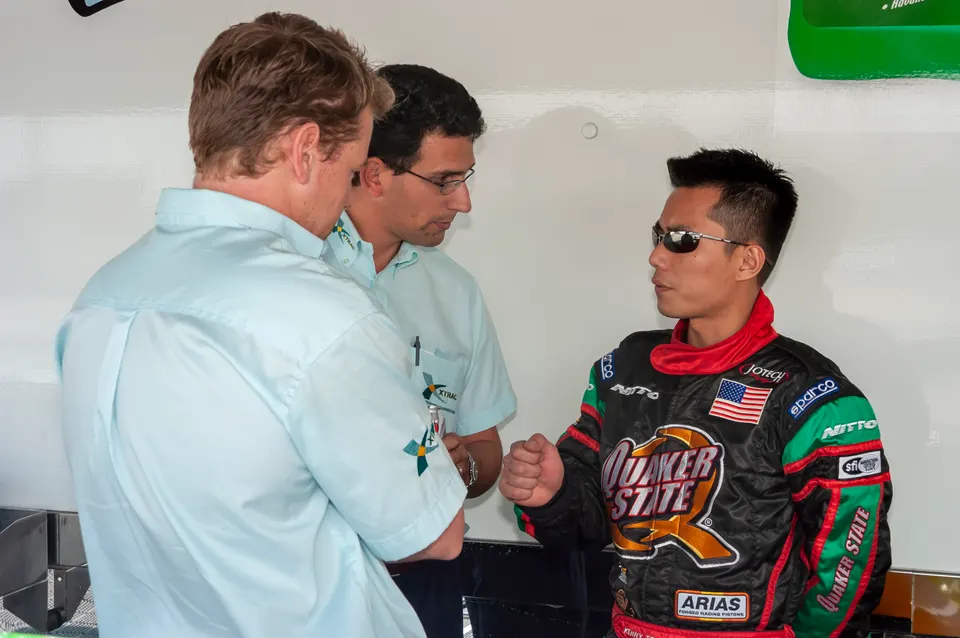 Three men, one in a racing suit, conversing in a garage or pit area at a motorsports event.