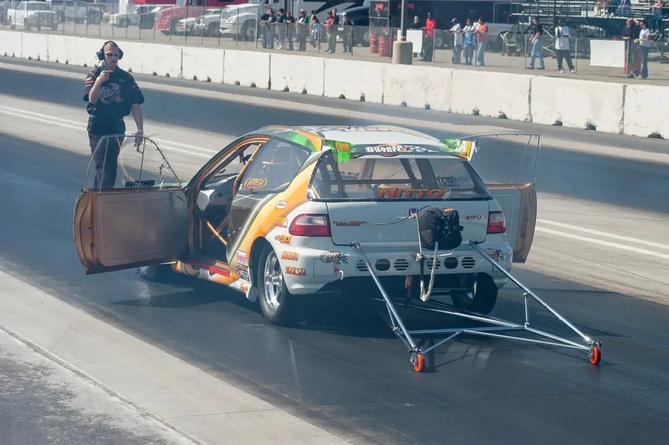 A white and orange Honda Civic drag car with a wheelie bar on a drag strip, with a person standing next to it during a burnout.