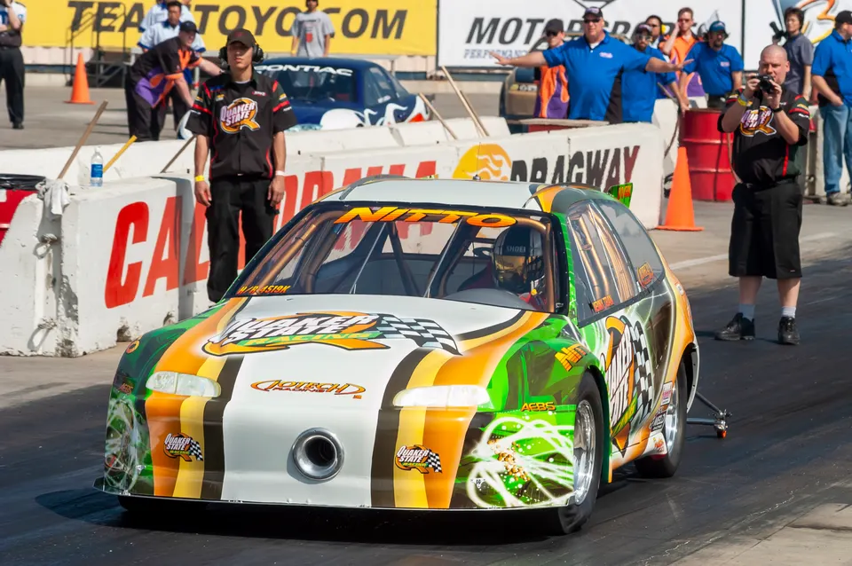 A highly modified drag race car with a custom green, orange, and white livery at the starting line of a drag strip.