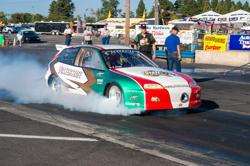 A green, white, and red Honda Civic race car performs a burnout on a drag strip, with smoke billowing from its tires.