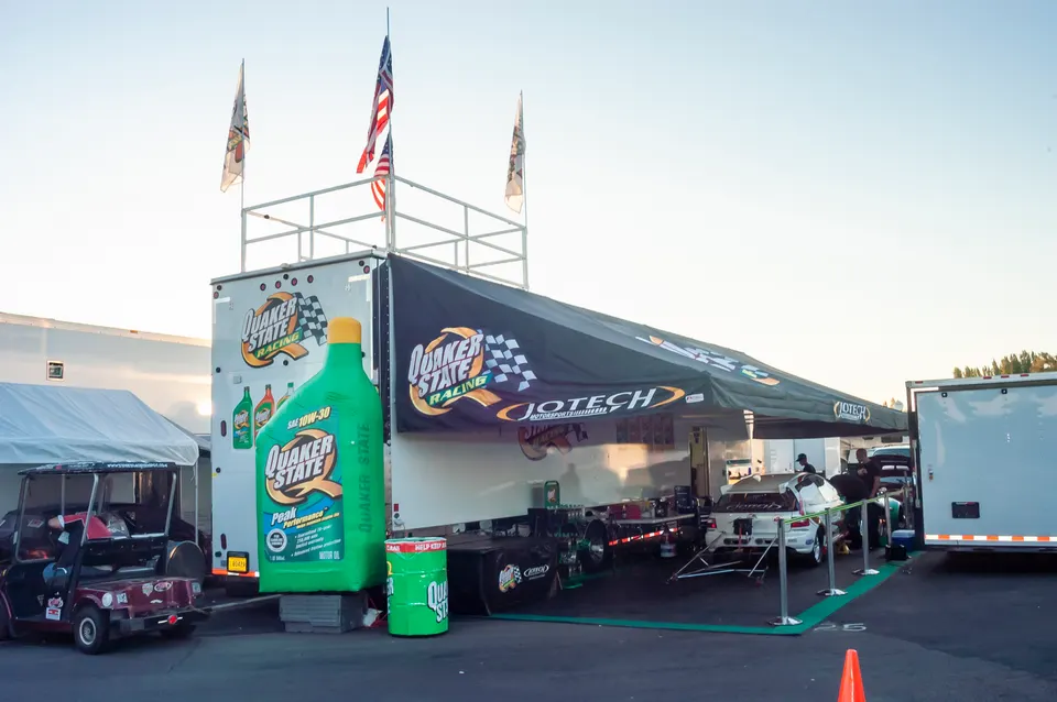 Quaker State Racing team trailer and drag car in the pit area at a race track event.