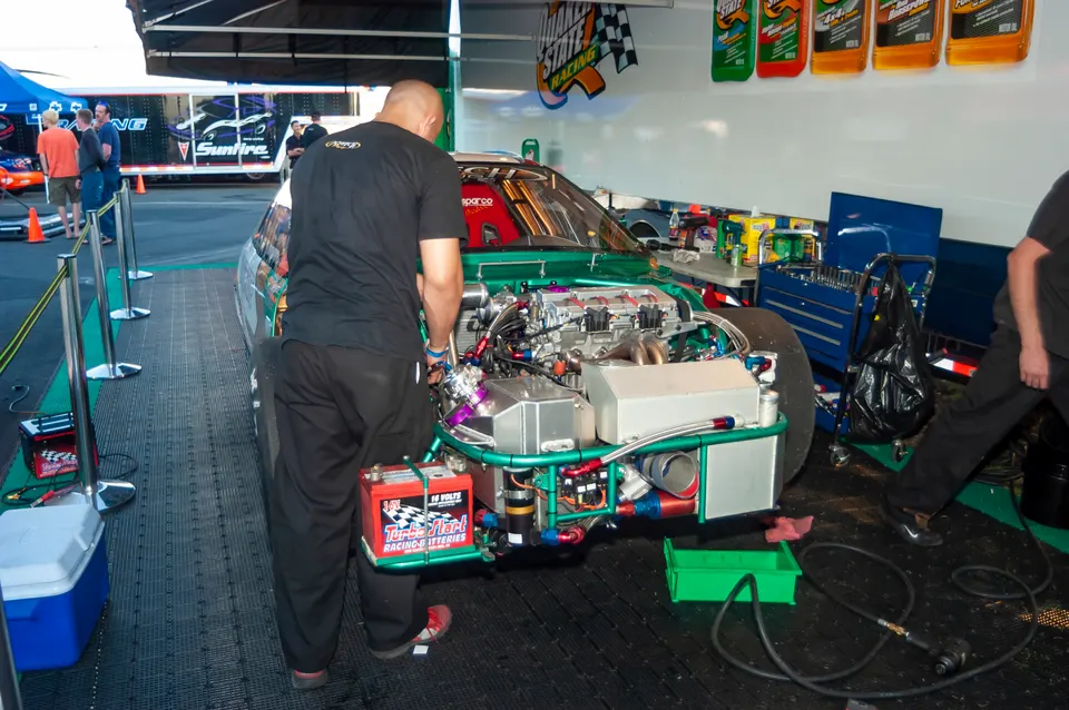 A mechanic works on the exposed engine of a green race car in a pit area, with tools and equipment nearby.
