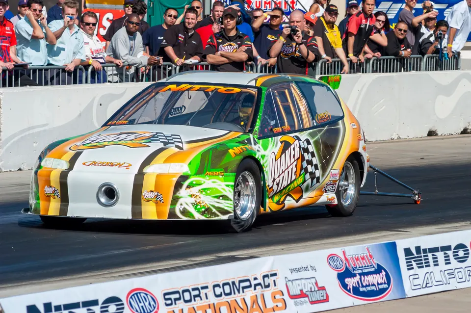 Highly modified Honda Civic drag car with Quaker State and Nitto livery on a drag strip with spectators in the background.