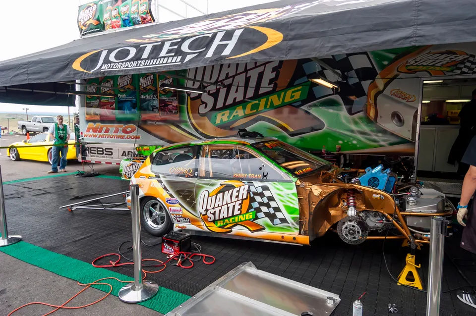 A Jotech Motorsports drag racing car with its front end exposed, showing suspension and brakes, under a Quaker State Racing tent.