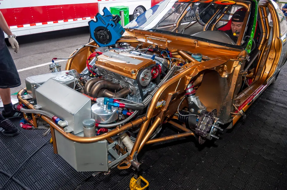 Highly modified gold drag car with exposed engine bay, custom chassis, and performance exhaust headers in a shop.