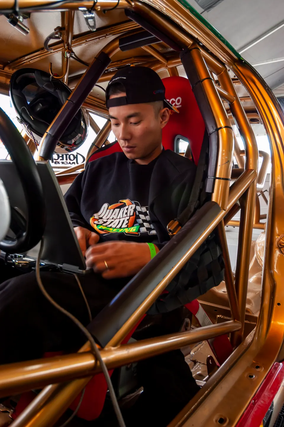 Driver in a race car interior with a gold roll cage, red racing seat, and safety harnesses, wearing a Quaker State Racing sweatshirt.