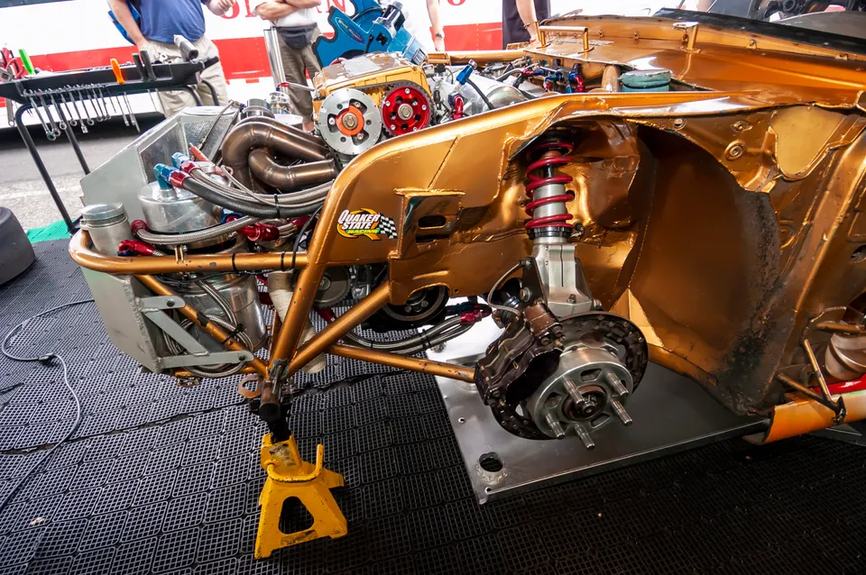 Front corner of a gold race car in a shop, showing exposed engine bay, custom suspension, and high-performance brakes.